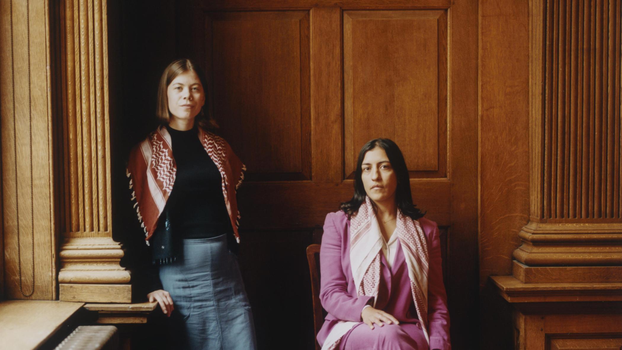 Two women one on chair one standing by window 